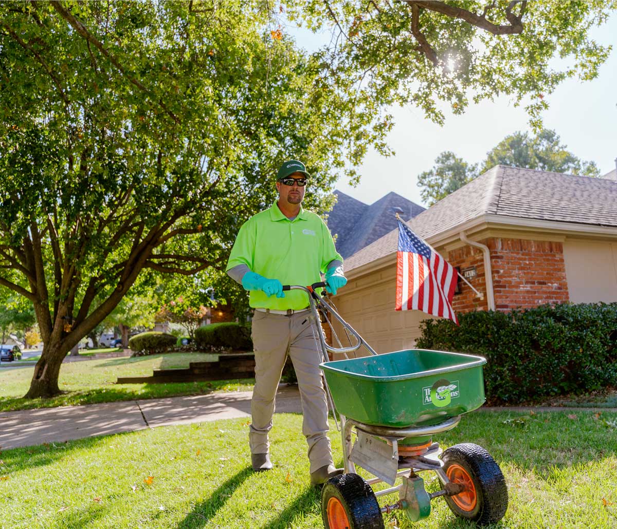 A man in a Green Group uniform standing in front of a green lawn with a fertilizer spreader
